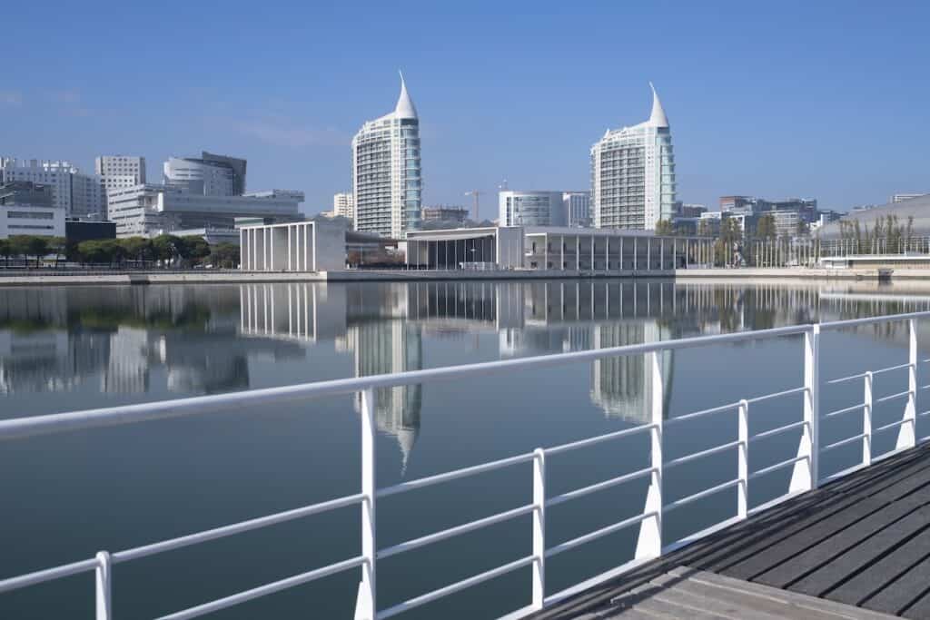 Modern waterfront view of Parque das Nações in Lisbon, Portugal, with twin sail-shaped towers, contemporary architecture, and clear reflections in the Tagus River under a bright blue sky.