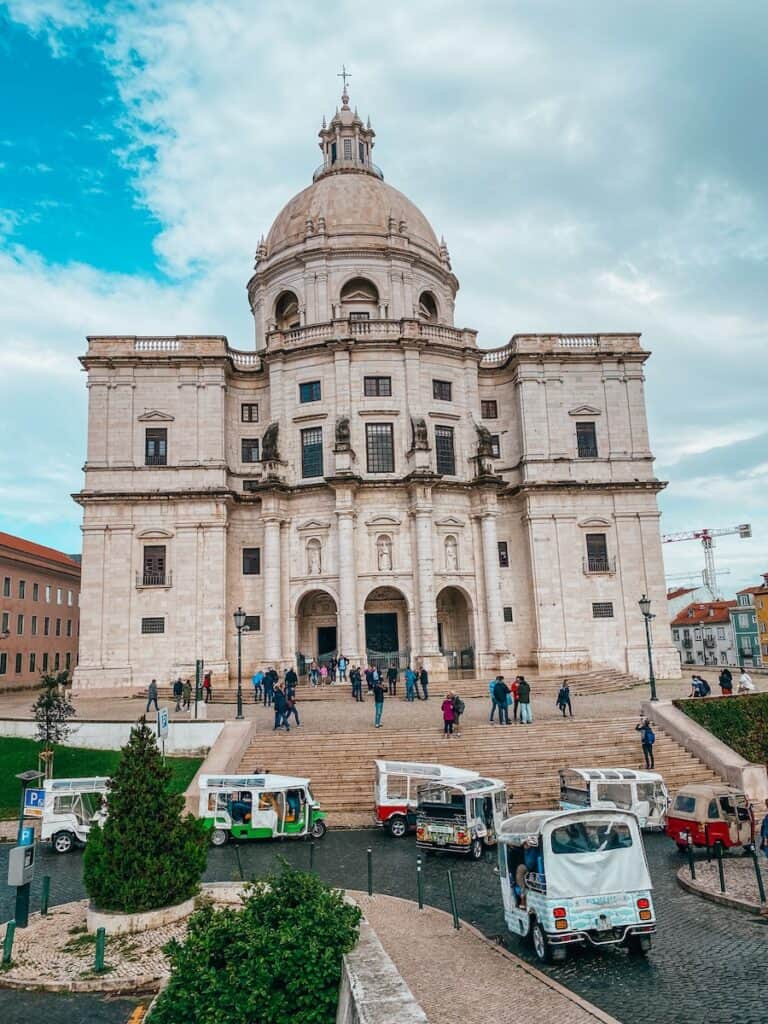 Front view of the National Pantheon in Lisbon, Portugal, with its grand baroque architecture and domed roof under a partly cloudy sky. A group of people stands on the wide stone staircase leading up to the entrance, while several colorful tuk-tuks are parked in the foreground.