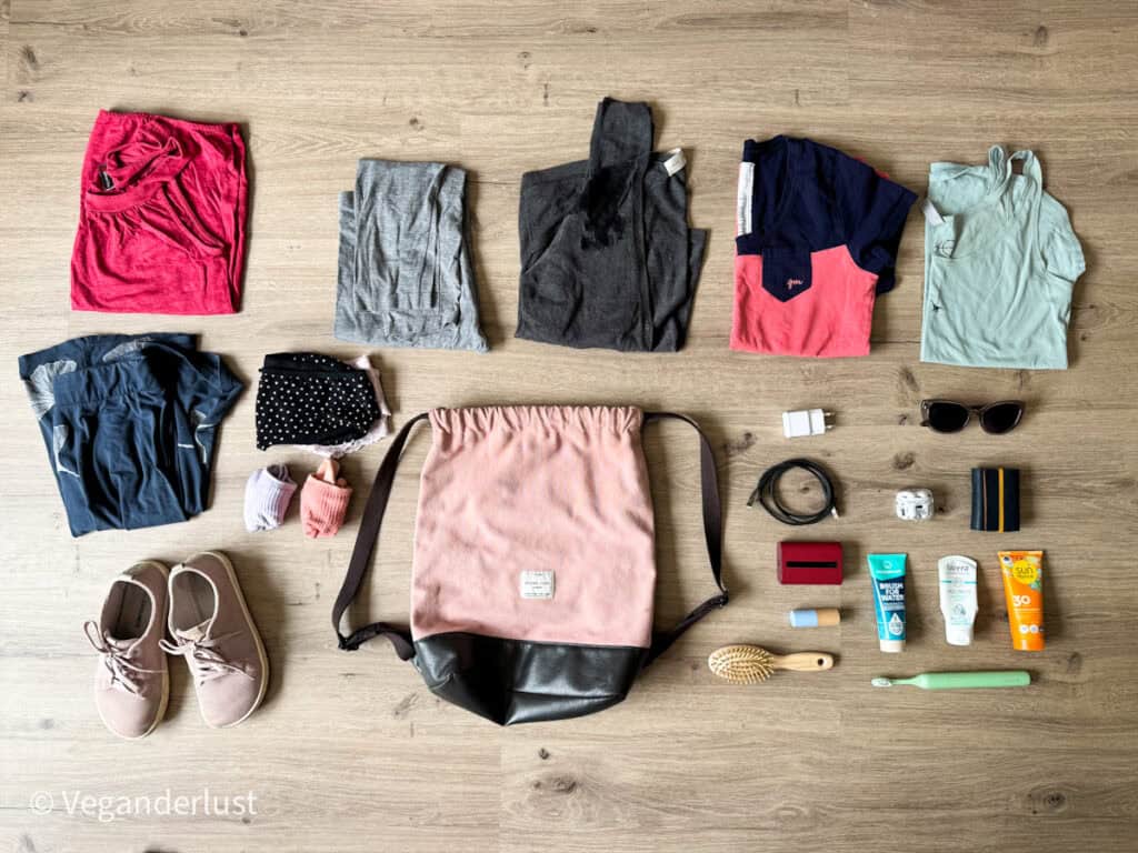 Flat lay of a minimalist travel packing list arranged beside a pink drawstring backpack, featuring lightweight clothing in soft colors, socks, underwear, casual shoes, sunglasses, chargers, skincare, sunscreen, toothpaste, and an electric toothbrush. The organized layout supports travel with only a personal item-sized bag.