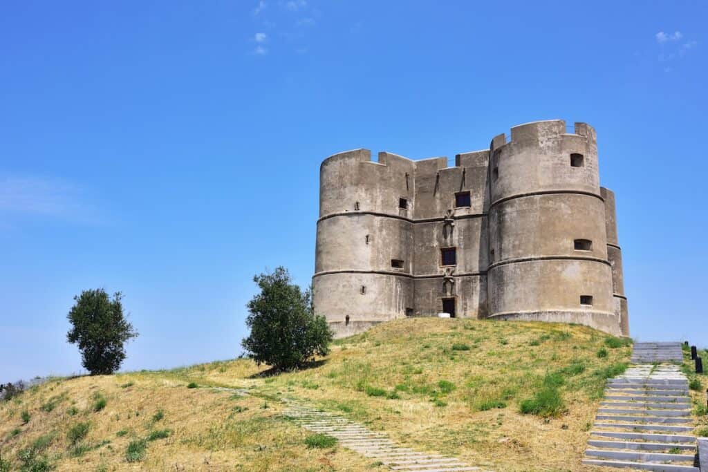 A solid, cylindrical fortress with rounded towers stands on a dry grassy hilltop beneath a clear blue sky, flanked by two sparse trees.