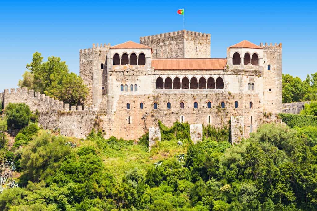 A large stone castle with red-tiled roofs and arched balconies flying the Portuguese flag, nestled among dense greenery on a bright sunny day.