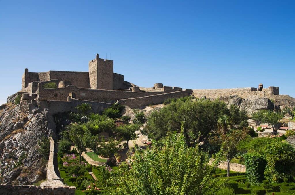 A mountaintop fortress with thick stone walls and towers overlooks a manicured garden filled with trees and flowers, under a clear blue sky.