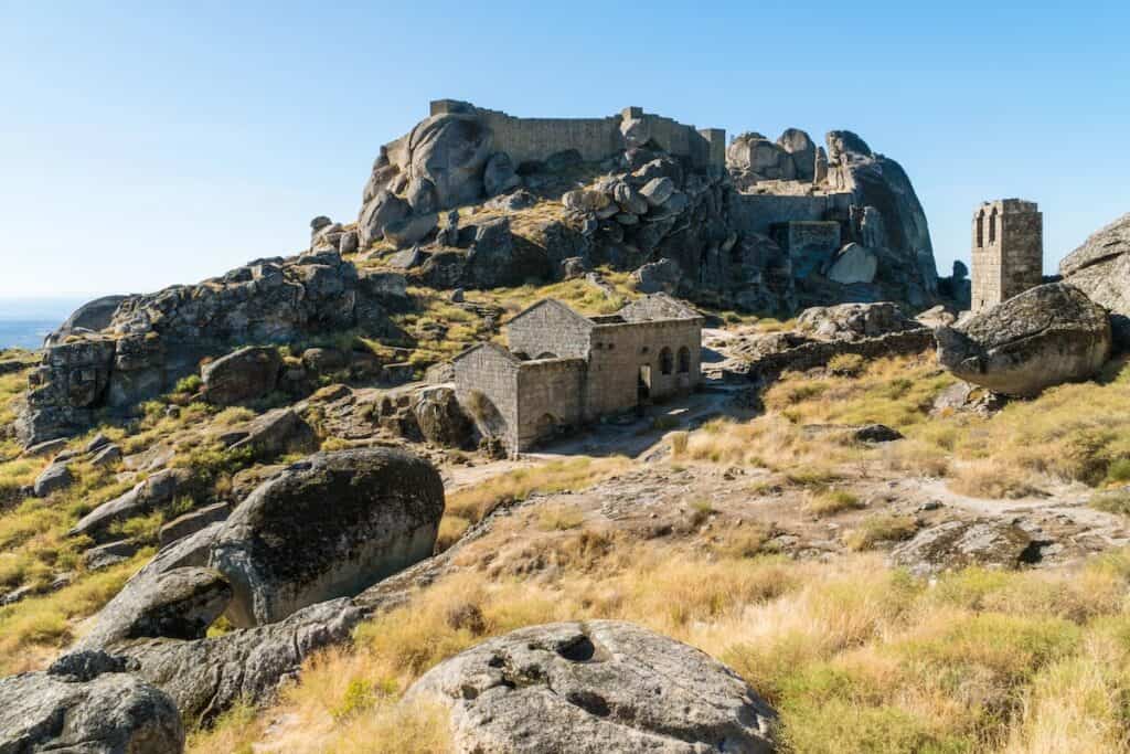 Ancient stone ruins and castle walls sit dramatically among massive boulders and dry grasses on a rugged mountaintop landscape.