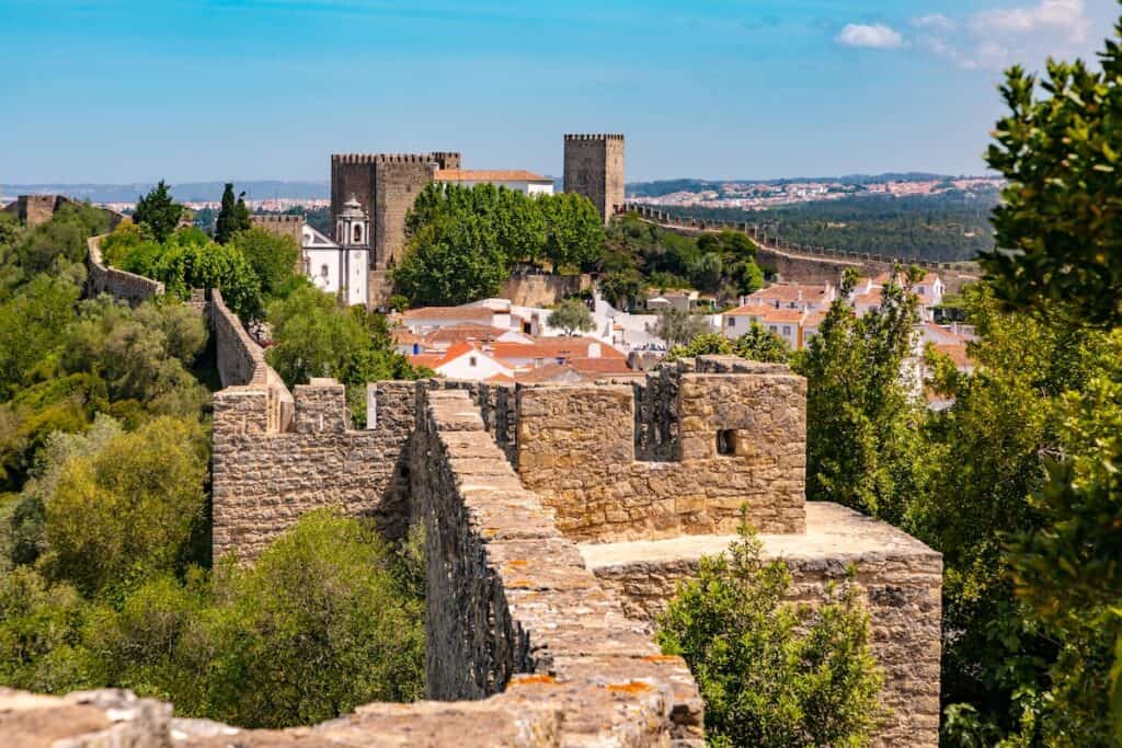 Stone castle walls and towers encircle a village with white and orange-roofed homes, framed by lush trees and a backdrop of distant hills.