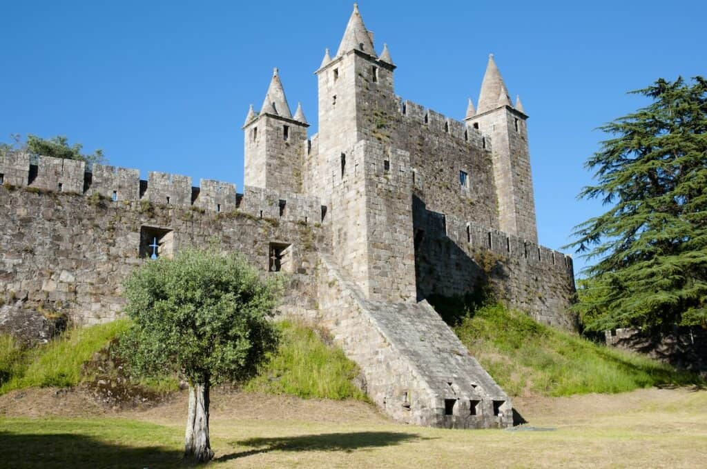 A well-preserved stone castle with pointed turrets and crenellated walls stands behind a single tree, set against a vibrant blue sky.