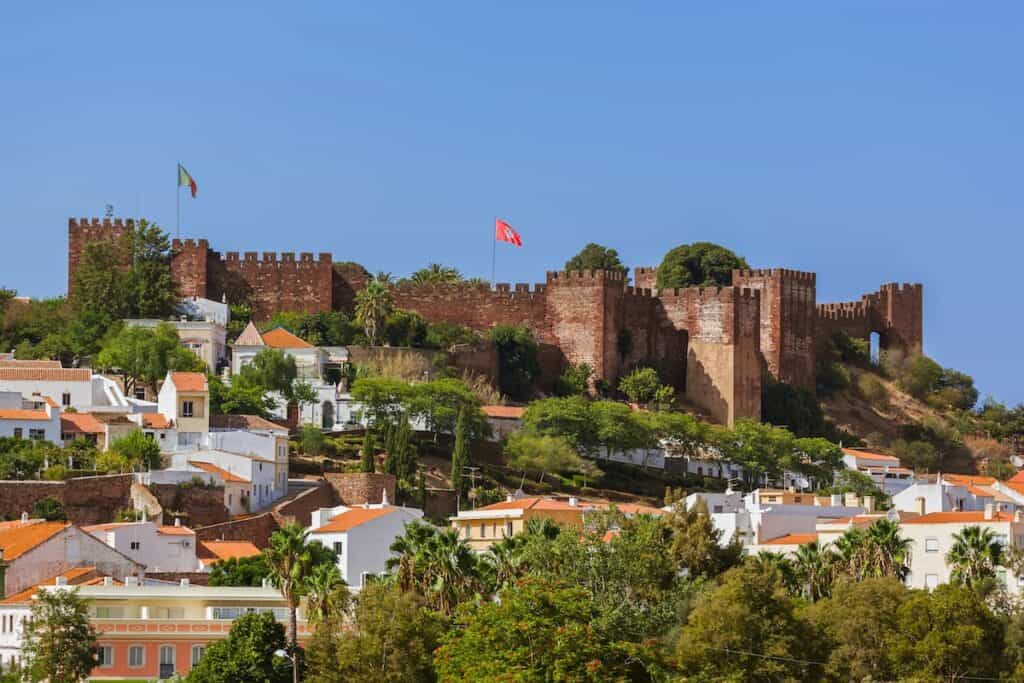 A red sandstone castle with crenellated walls and towers rises above a hillside town of white houses with orange roofs and palm trees.