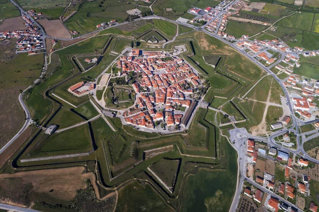 An aerial shot of a fortified town with perfectly preserved star-shaped bastions and red-roofed buildings, surrounded by green fields and modern roads.