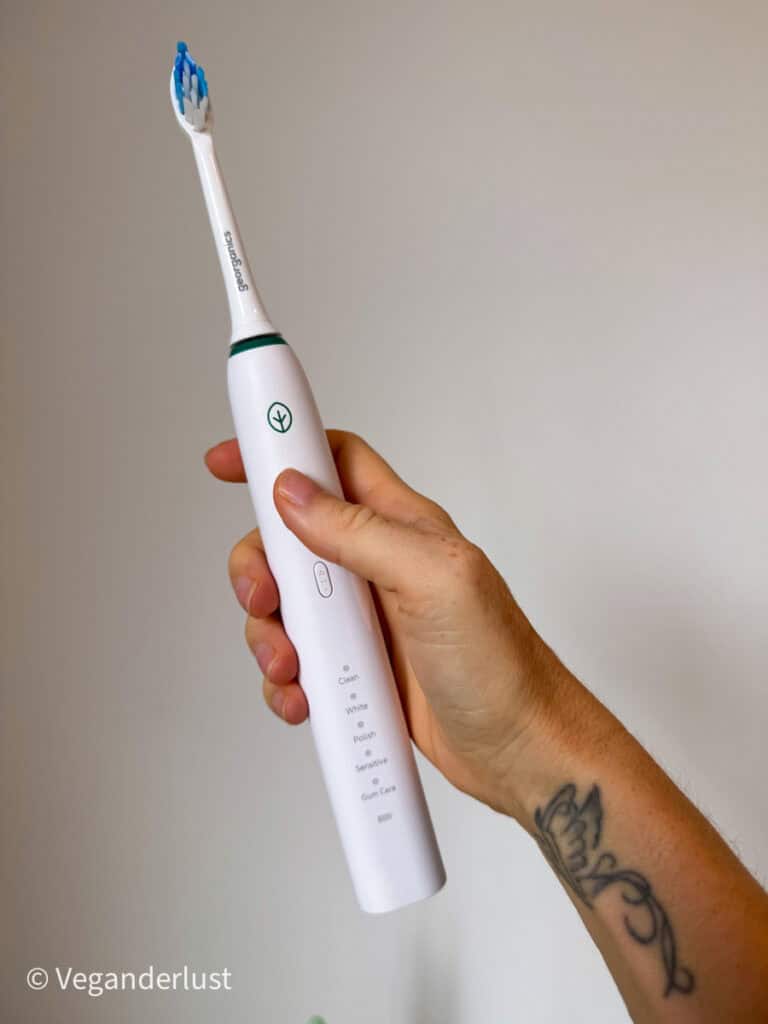 A hand holding a white Georganics electric toothbrush upright, showing its various brushing modes like Clean, White, Polish, Sensitive, and Gum Care. A leafy logo and tattooed wrist are also visible.