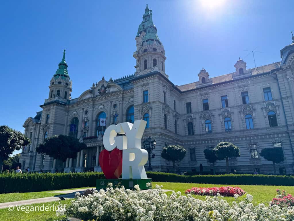 Grand neo-baroque city hall of Győr with ornate detailing, green copper domes, and symmetrical towers. In front, large “GYŐR” letters with a red heart stand in a manicured garden filled with white and pink flowers.