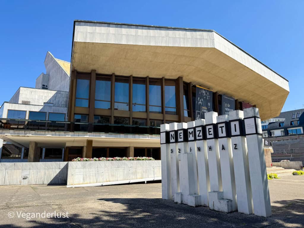 Brutalist-style building with angular lines and large glass windows, labeled "Nemzeti Színház" (National Theatre). The foreground includes a matching concrete sign with black lettering and shadows falling across the pavement.