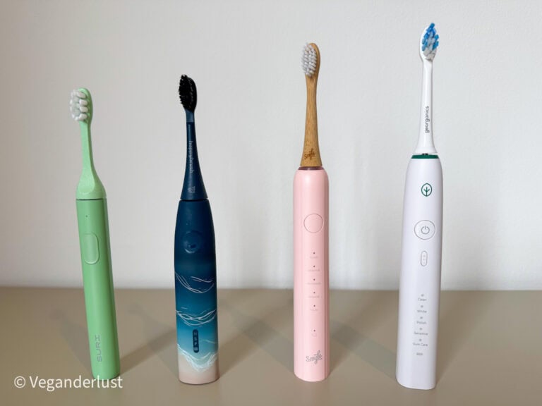 Four different electric toothbrushes standing upright on a flat surface. From left to right: a green Suri toothbrush, a blue HappyBrush toothbrush, a pink Smyle toothbrush, and a white Georganics toothbrush.