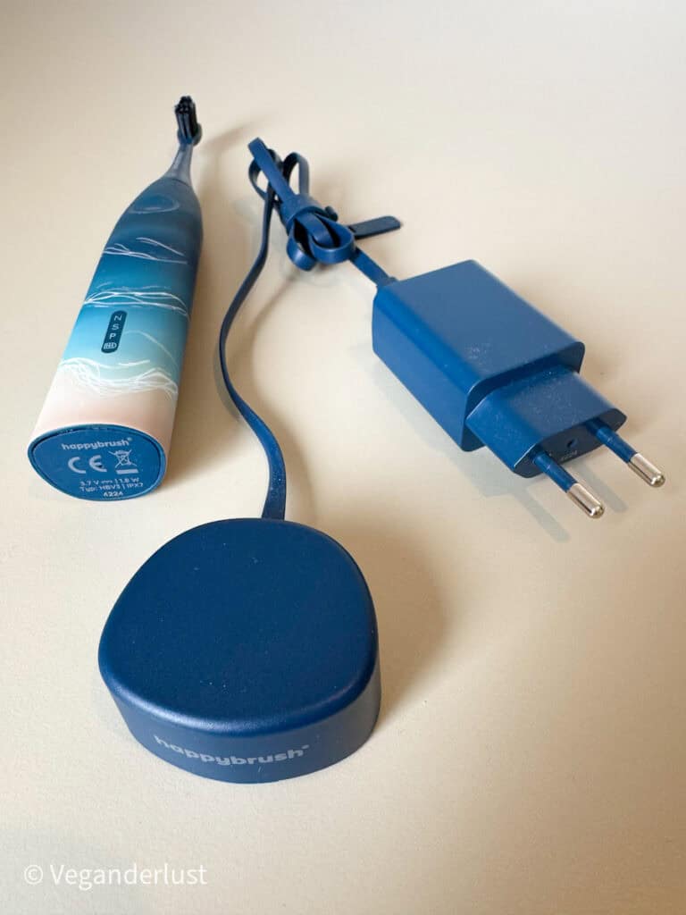 A blue HappyBrush electric toothbrush laying on a flat surface with its matching blue charging cable and plug. The toothbrush has a wave-like pattern on the handle and a black brush head.