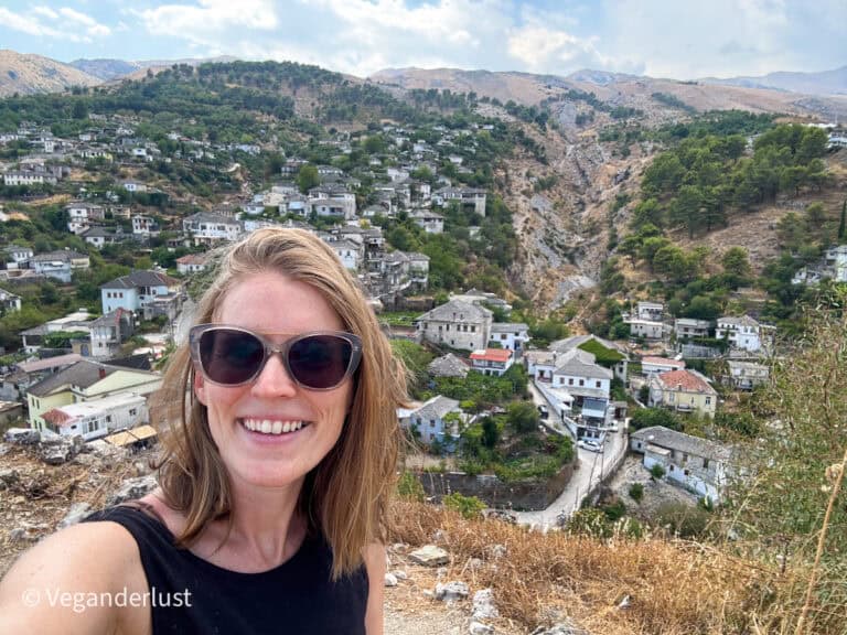 Tina in a selfie with a scenic hillside village in the background, filled with white stone houses and surrounded by green trees and rugged mountains. She wears sunglasses and a black tank top, with the watermark “@Veganderlust” in the corner.
