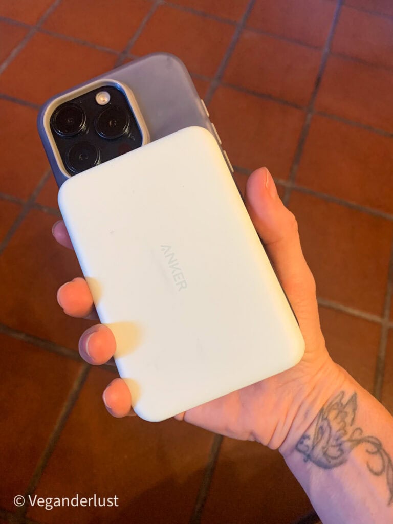 A hand holding a white Anker power bank magnetically attached to the back of a smartphone with three rear cameras. The background features red tiled flooring and part of a forearm tattoo is visible.