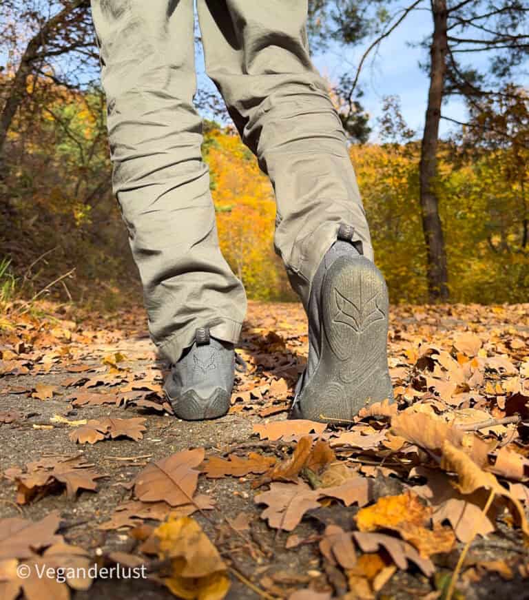 A close-up shot from behind shows someone walking through a leaf-covered path in the forest, wearing black Wildling barefoot shoes and olive green pants. Fallen brown oak leaves line the sunlit trail.