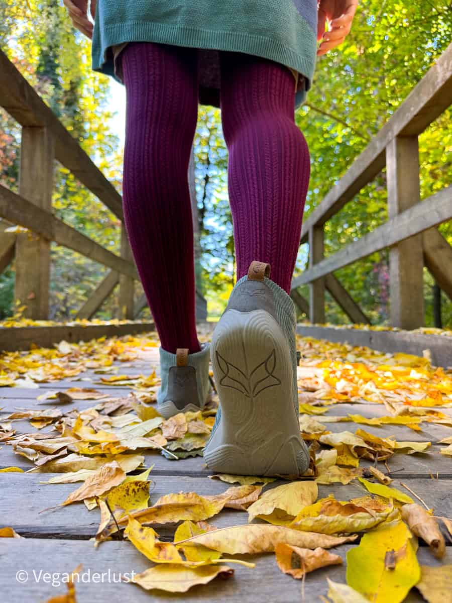A person walks across a wooden bridge covered in crisp yellow autumn leaves, wearing light-colored Wildling barefoot shoes and deep burgundy textured tights. The scene is framed by vibrant fall foliage in a wooded park.