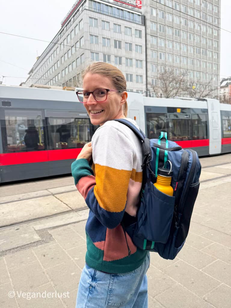 Tina wearing the BAGSMART Blast Elite 22L backpack with a yellow bottle in the side pocket, standing at a tram stop in an urban setting.