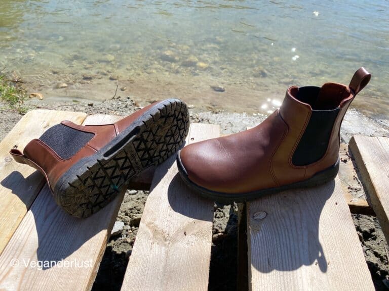 A pair of brown vegan Chelsea boots in new condition, with clean uppers and treaded soles, displayed on wooden planks by a clear lake.