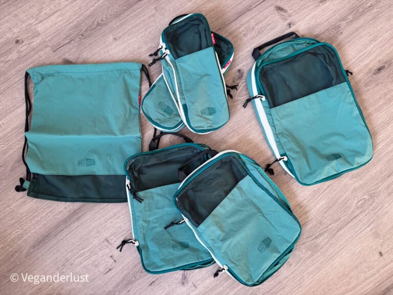 A set of teal packing cubes and a matching drawstring laundry bag are laid out on a wooden floor. The cubes feature black mesh panels, white zippers, and the Bagsmart logo on the front. This image shows the full organization set included for travel packing.