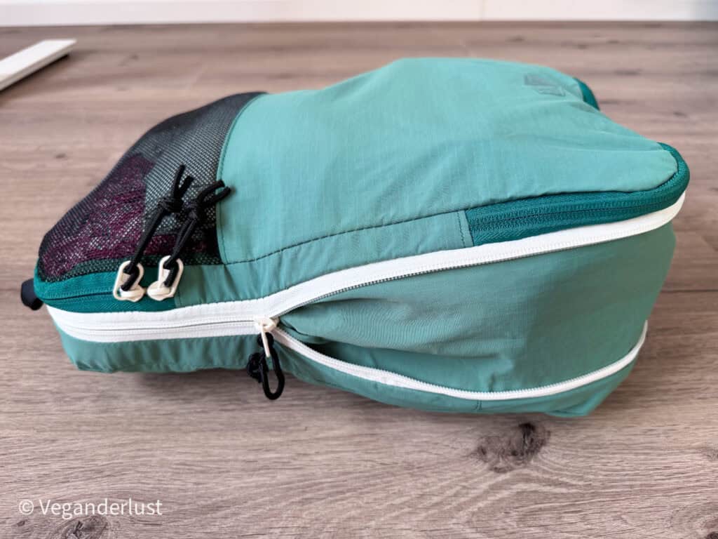 Side view of a teal compression packing cube with dual white zipper tracks running around the middle. The mesh panel and zipper pulls are visible along the top edge. This angle shows how the cube expands and compresses for adjustable packing space.