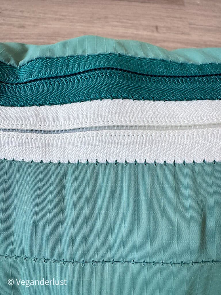 Extreme close up of the teal fabric and zipper stitching on a packing cube. The image highlights the white zipper track, green zipper tape, and reinforced seams. This detail emphasizes construction quality and material texture.