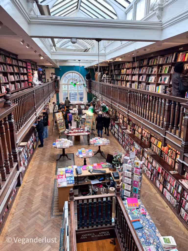 Grand interior of a historic bookstore with a glass ceiling skylight and two levels of wooden balconies lined with books. Customers browse tables and shelves beneath green reading lamps and a large arched window.