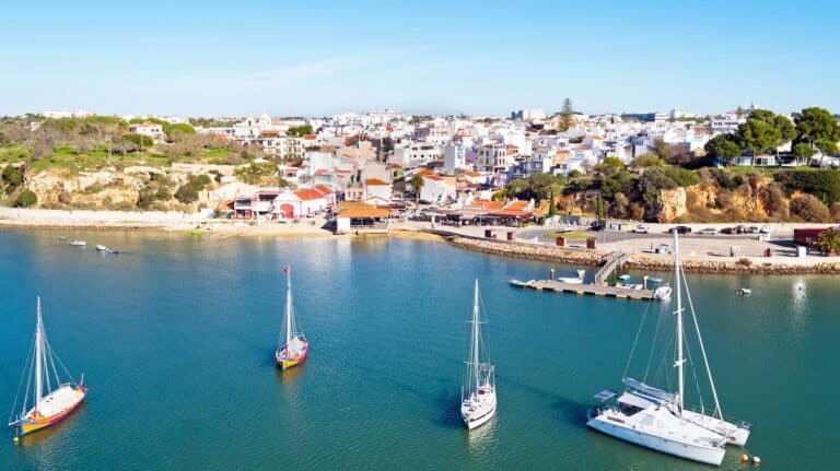 Aerial view of a coastal town in Portugal with white buildings and terracotta roofs lining a calm turquoise harbor. Several sailboats and a catamaran float on the water near a small marina and waterfront promenade. The image captures a sunny Algarve seaside scene with boats anchored just offshore.