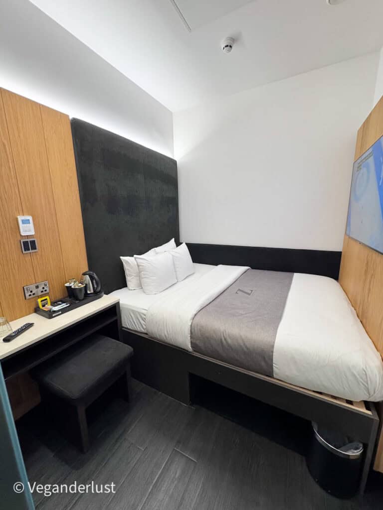 Compact modern hotel room with a double bed against a dark upholstered headboard and light wood paneling. A small desk with a stool and kettle sits beside the bed, highlighting the efficient use of space.