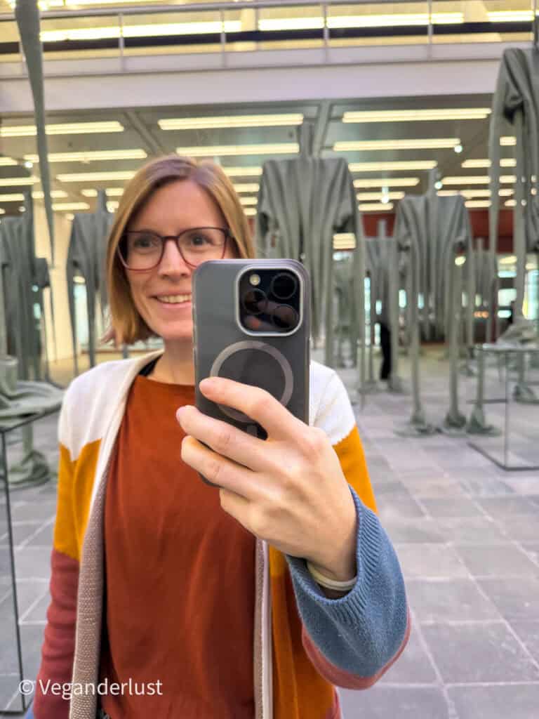 Mirror selfie of a woman holding a smartphone with a magnetic ring case inside a modern art installation. Tall sculptural figures fill the background under bright gallery lights. The photo shows the phone setup in use while exploring a museum space.
