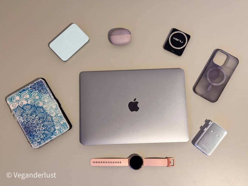 Flat lay of travel tech essentials arranged around a closed silver MacBook with Apple logo centered on a light surface. Items include a pink earbud charging case, a foldable magnetic phone stand, a clear phone case with magnetic ring, a compact Anker wall charger, a slim white power bank, a patterned passport holder, and a pink smartwatch band. The organized layout highlights portable gadgets and accessories for working while traveling.