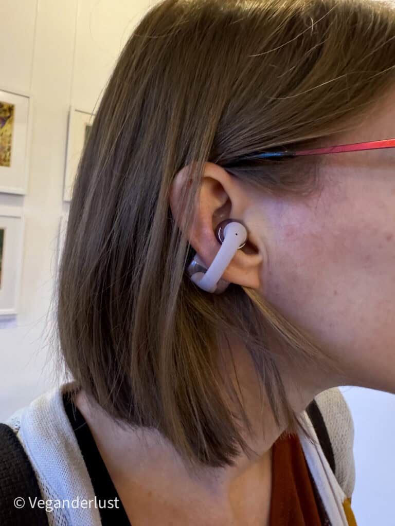 Close up of a woman wearing a wireless earbud hooked securely around her ear. Her short brown hair is tucked behind her ear and she wears red framed glasses. The image highlights how the earbud fits comfortably for everyday use.