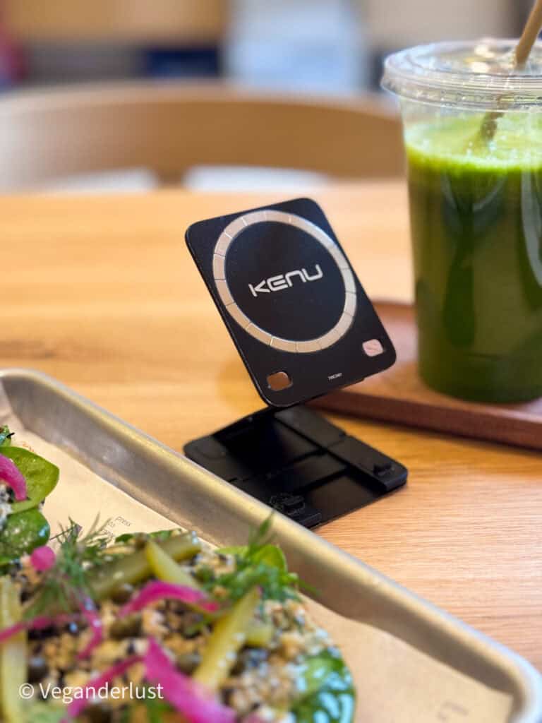 Foldable Kenu magnetic phone stand set up on a cafe table next to a green juice and salad. The circular magnetic ring is centered on the front panel. The scene shows how the stand props up a phone for hands free use while dining.