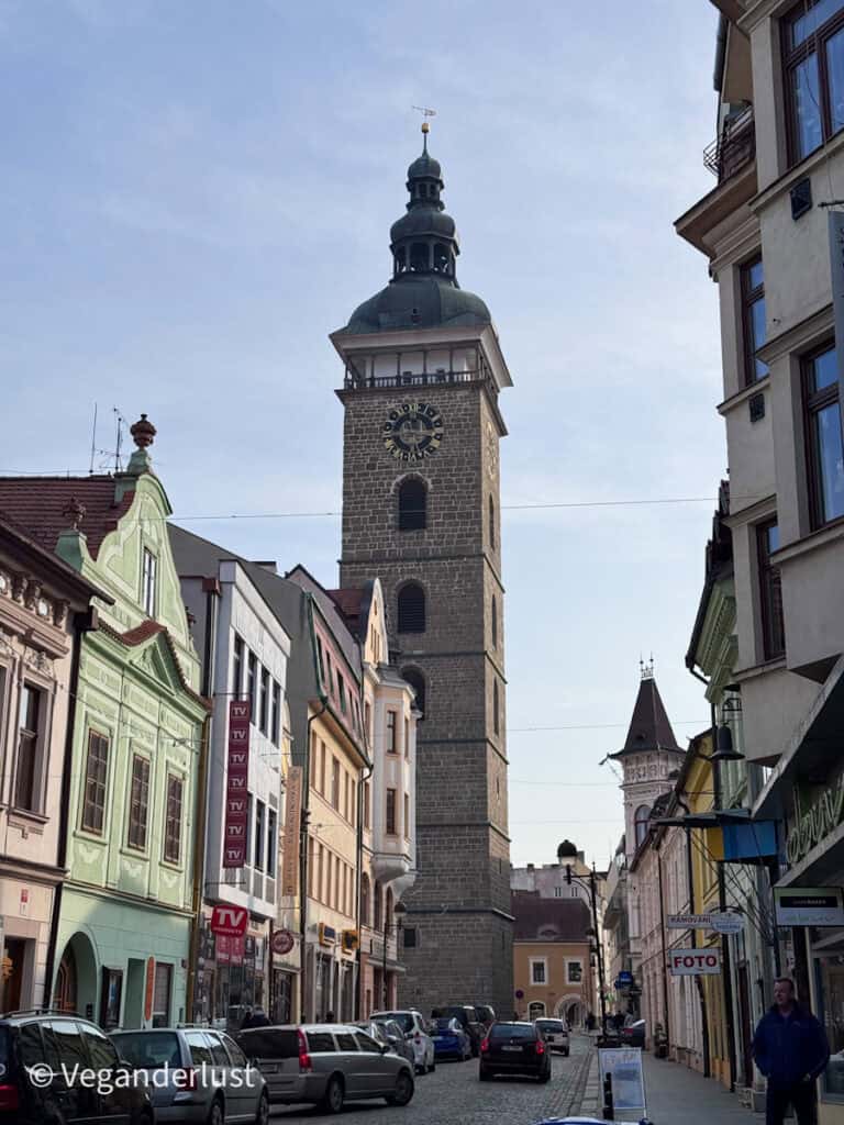 Historic street in České Budějovice lined with colorful buildings leading toward a tall stone clock tower with a dark domed top and large clock face. Parked cars and shop signs sit along the cobblestone road beneath the tower in the city center.