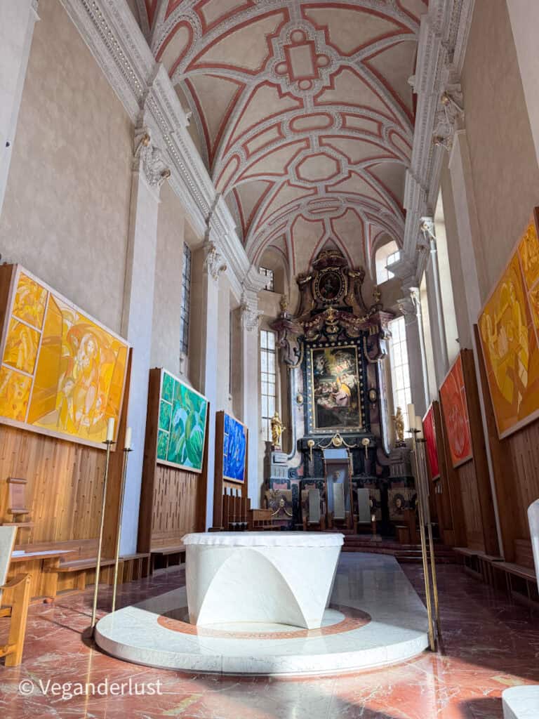 Interior of a church in České Budějovice with a white marble altar in the center aisle and a high vaulted ceiling painted with red decorative patterns. Large colorful modern religious paintings hang along the walls leading toward an ornate altar at the back.
