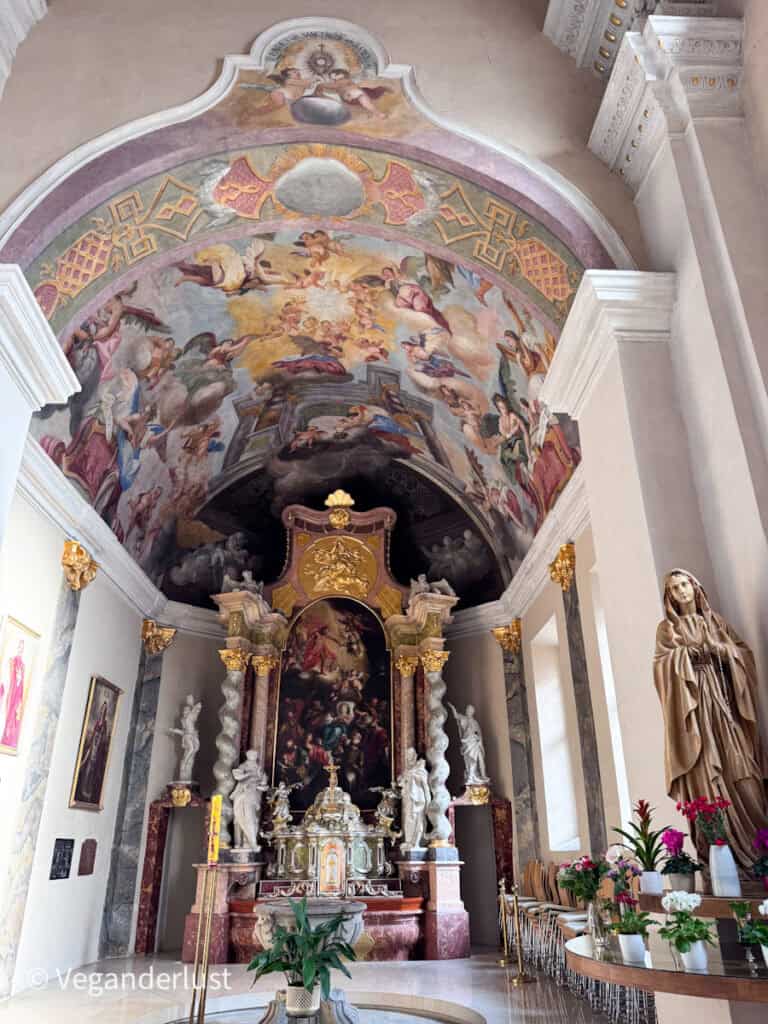 Ornate side chapel inside a church in České Budějovice with a richly decorated altar framed by marble columns and statues. A ceiling fresco filled with angels and clouds spreads above the altar while flowers and candles sit nearby.