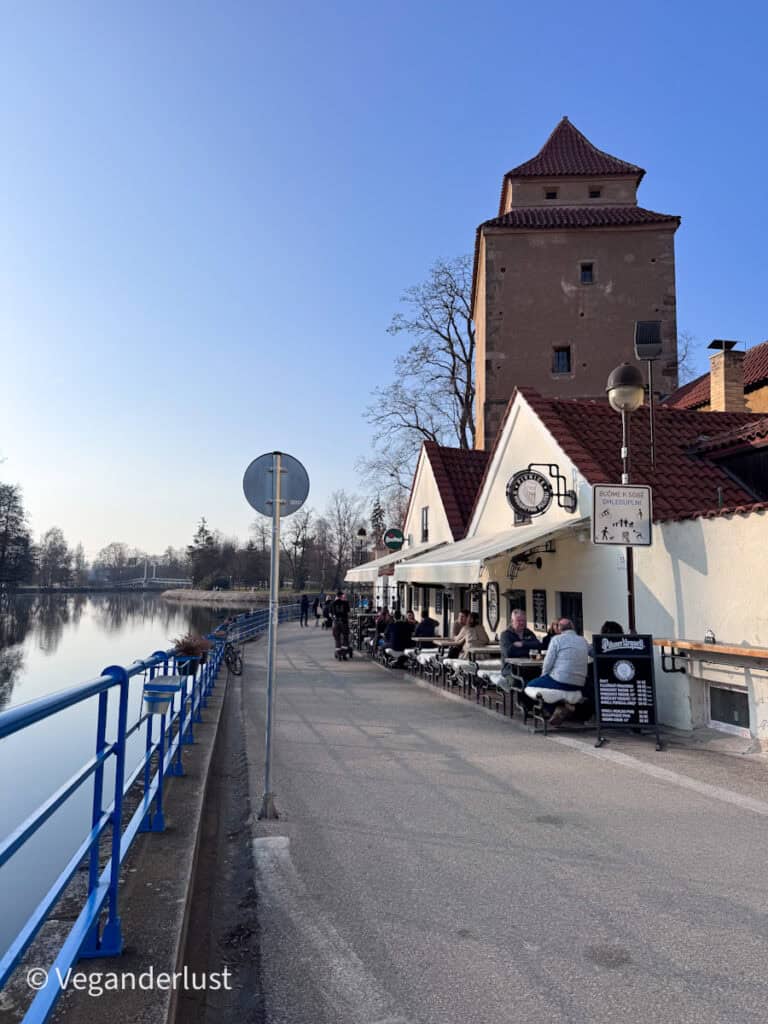 Riverside promenade in České Budějovice with outdoor restaurant seating beside the water and a historic stone tower behind the buildings. People sit at tables along the path enjoying the view of the river.
