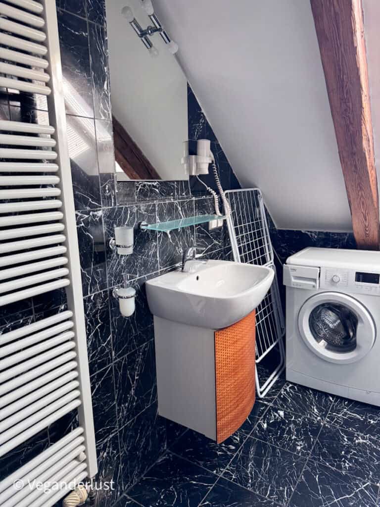 Compact bathroom with dark marble patterned tiles a small sink wall mirror and a washing machine tucked beneath the sloped ceiling.