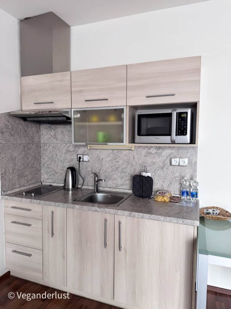 Small kitchenette with light wood cabinets a sink stovetop electric kettle and microwave arranged along a compact counter.