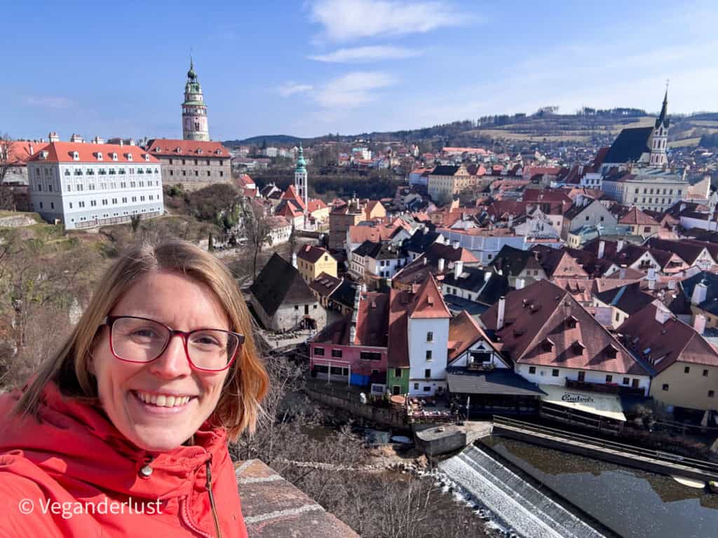 Tina in a red jacket smiles at the camera while standing at a viewpoint overlooking the red rooftops, river bend, and Cesky Krumlov Castle tower. Enjoying panoramic viewpoints like this is one of the most memorable things to do in Cesky Krumlov.