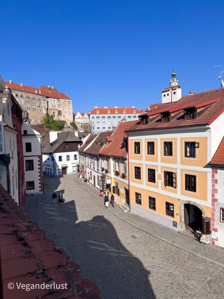 Cobblestone street lined with pastel historic buildings leads toward Cesky Krumlov Castle rising above the town under a bright blue sky. Walking through the old town streets offers scenic views around every corner while exploring things to do in Cesky Krumlov.