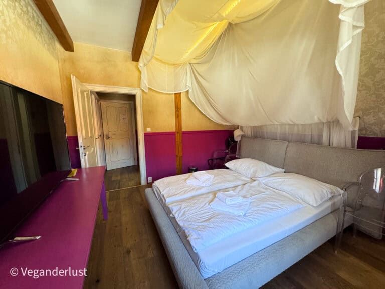 Cozy hotel room with a large bed under a draped canopy, pink accent walls, wooden floors, and folded towels placed on the bed. Unique boutique accommodations add to the experience of staying overnight while discovering things to do in Cesky Krumlov.