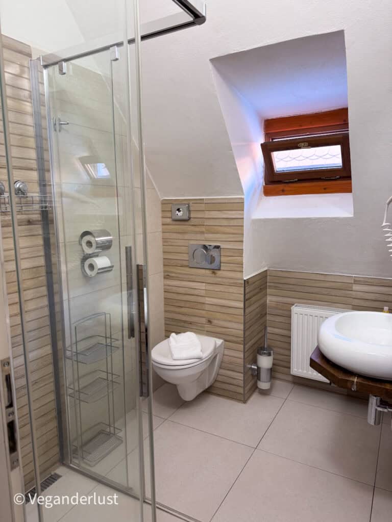 Modern hotel bathroom with glass shower, wall mounted toilet, small window, and round sink set against beige tile walls. Comfortable amenities like this help make a stay convenient while exploring things to do in Cesky Krumlov.