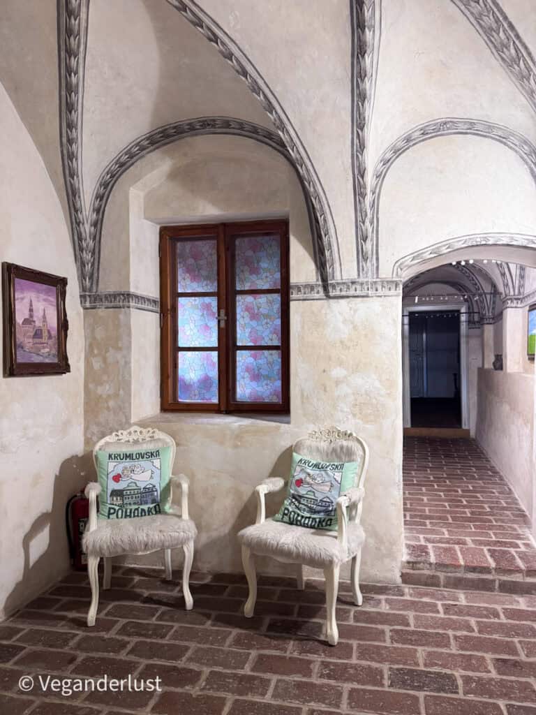 Historic hallway with vaulted ceilings, decorative painted arches, and two white chairs with pillows reading "KRUMLOVSKÁ POHÁDKA." The elegant interior reflects the historic architecture found in many buildings around Cesky Krumlov.