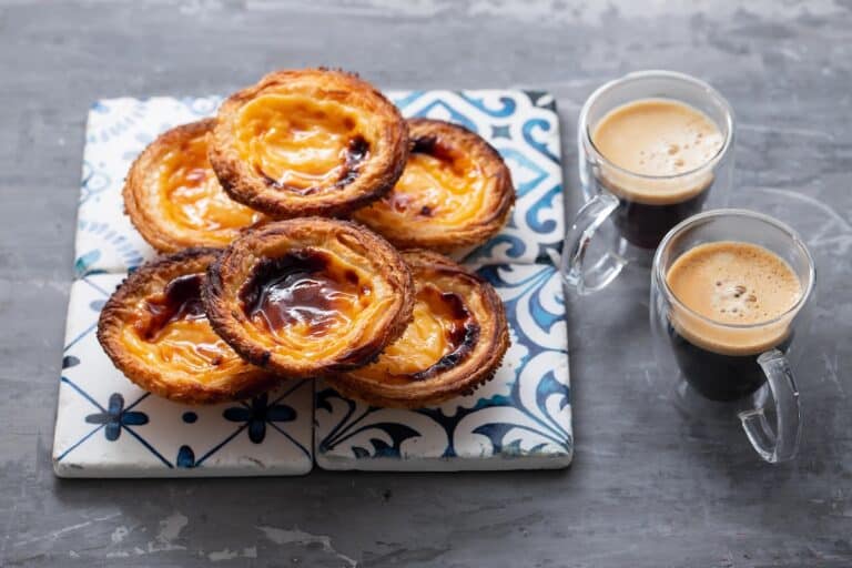 Golden Portuguese custard tarts with caramelized tops arranged on patterned tiles beside two small glass cups of espresso with crema on a gray table.