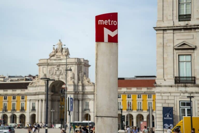 Red metro sign with a white "M" stands in a city square with a historic arch and yellow buildings in the background as people walk through the area.