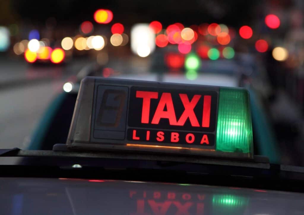 Illuminated taxi sign reading "TAXI LISBOA" on top of a car at night with blurred city lights and traffic in the background.