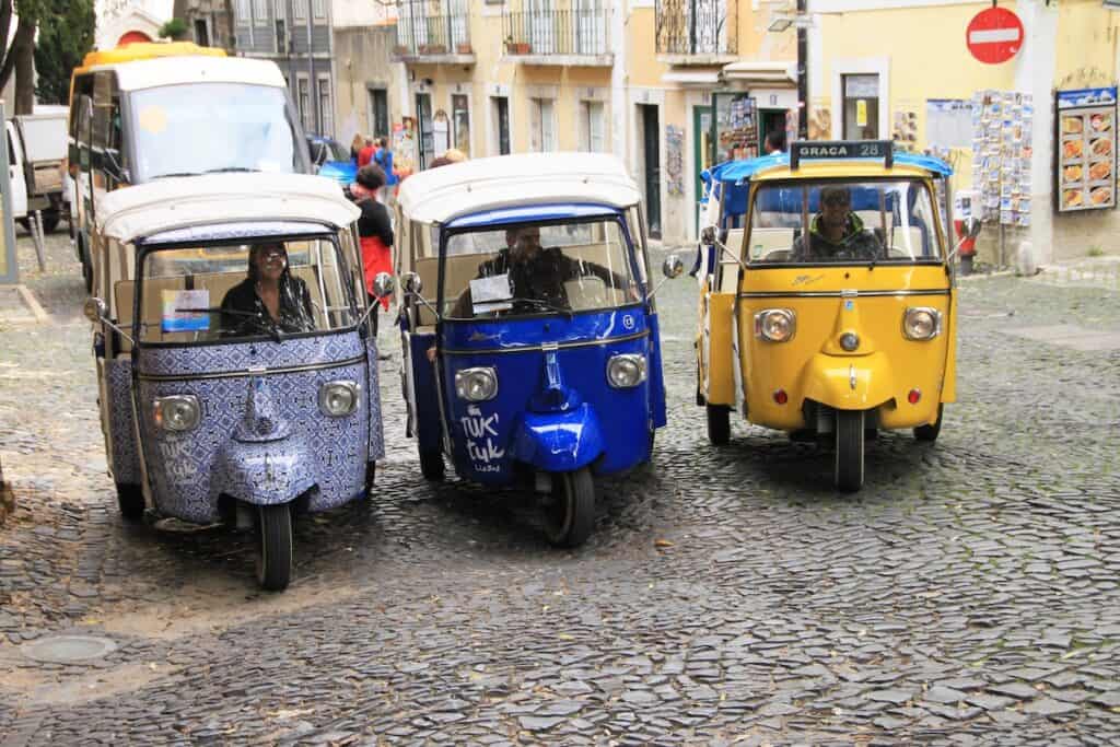 Three colorful tuk tuk taxis drive side by side on a cobblestone street lined with pastel buildings, with drivers visible inside and a sign on one reading "Graça 28".