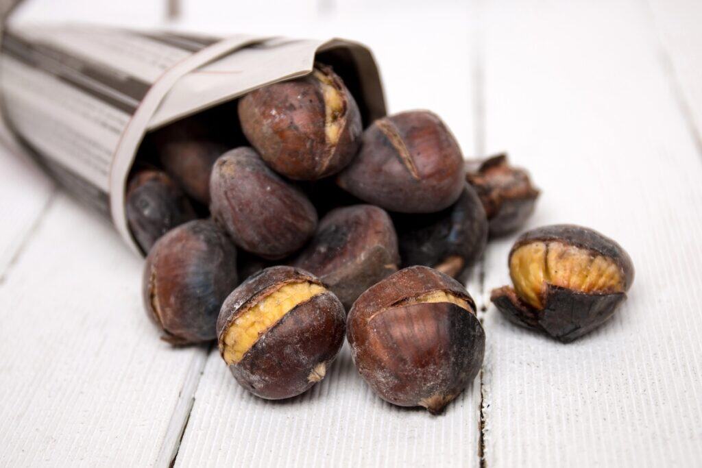 Roasted chestnuts spilling out of a paper cone onto a white wooden surface, with several split open to reveal the soft yellow interior.