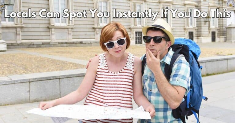 Two tourists wearing sunglasses and casual summer clothes stand in front of a historic building while looking at a paper map, with text above them reading "Locals Can Spot You Instantly If You Do This".