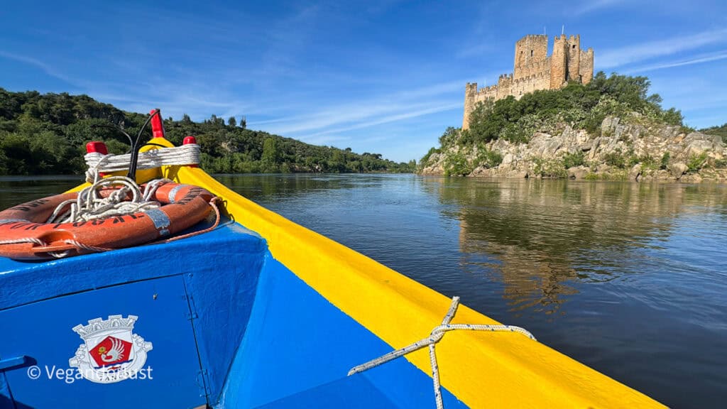 View from a small boat with bright yellow and blue edges looking toward a stone castle on a rocky hill beside a calm river. A life ring and rope sit on the boat while the castle reflects in the water under a clear blue sky.